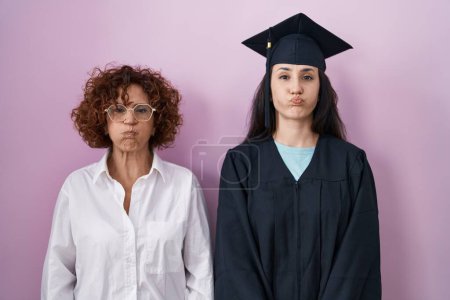 Photo for Hispanic mother and daughter wearing graduation cap and ceremony robe puffing cheeks with funny face. mouth inflated with air, crazy expression. - Royalty Free Image