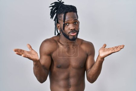 Téléchargez les photos : Homme africain avec dreadlocks debout torse nu sur fond isolé expression désemparée et confus avec les bras et les mains levées. concept de doute. - en image libre de droit