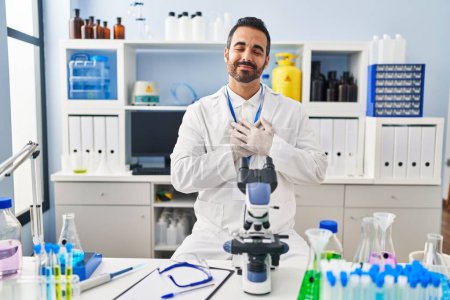 Téléchargez les photos : Jeune homme hispanique avec barbe travaillant au laboratoire scientifique souriant avec les mains sur la poitrine avec les yeux fermés et geste reconnaissant sur le visage. concept de santé. - en image libre de droit