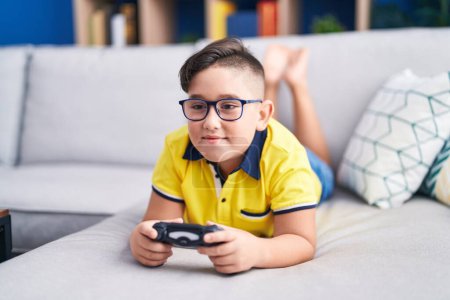 Téléchargez les photos : Jeune enfant hispanique jouant contrôleur de tenue de jeu vidéo sur le canapé clin d'oeil en regardant la caméra avec une expression sexy, gai et heureux visage. - en image libre de droit