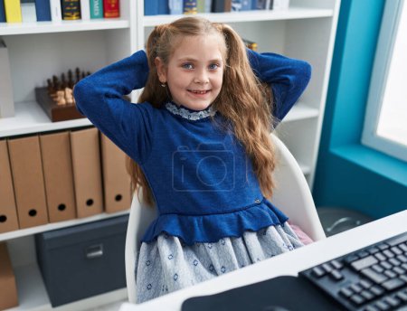 Foto de Adorable estudiante rubia chica usando la computadora descansando con las manos en la cabeza en el aula - Imagen libre de derechos