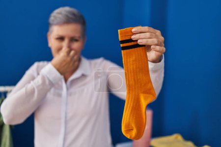 Téléchargez les photos : Femme d'âge moyen souriant confiant odeur chaussette sale à la buanderie - en image libre de droit