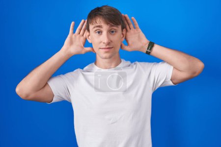 Photo for Caucasian blond man standing over blue background trying to hear both hands on ear gesture, curious for gossip. hearing problem, deaf - Royalty Free Image