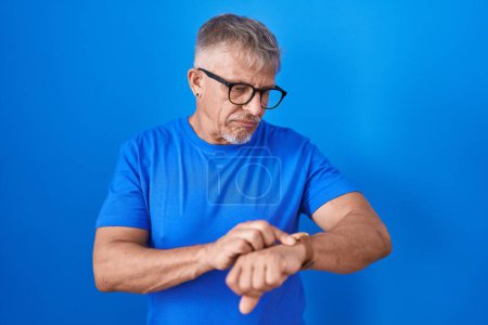 Foto de Hombre hispano con el pelo gris de pie sobre fondo azul comprobar la hora en el reloj de pulsera, relajado y seguro - Imagen libre de derechos