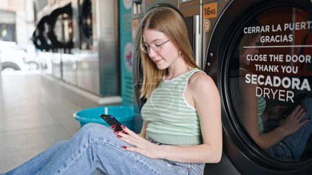 Téléchargez les photos : Jeune femme blonde utilisant un smartphone attendant la machine à laver à la buanderie - en image libre de droit
