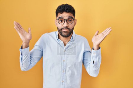 Photo for Hispanic man with beard standing over yellow background clueless and confused expression with arms and hands raised. doubt concept. - Royalty Free Image