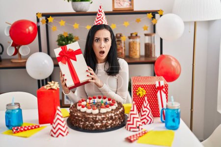 Téléchargez les photos : Femme hispanique célébrant l'anniversaire avec gâteau tenant présent dans le visage de choc, l'air sceptique et sarcastique, surpris avec la bouche ouverte - en image libre de droit