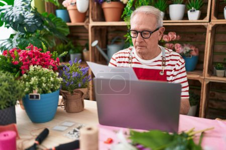 Foto de Middle age grey-haired man florist using laptop reading document at flower shop - Imagen libre de derechos