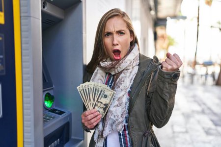 Téléchargez les photos : Jeune femme blonde tenant des billets de dollars de la machine à guichet automatique ennuyé et frustré criant de colère, criant fou de colère et la main levée - en image libre de droit