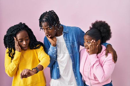 Foto de Grupo de tres jóvenes negros de pie juntos sobre fondo rosa mirando el tiempo reloj preocupado, con miedo de llegar tarde - Imagen libre de derechos