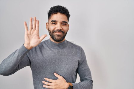 Foto de Hombre hispano con barba de pie sobre fondo blanco renunciando a decir hola feliz y sonriente, gesto de bienvenida amistoso - Imagen libre de derechos