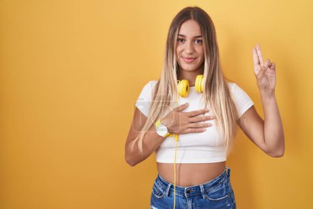 Téléchargez les photos : Jeune femme blonde debout sur fond jaune portant des écouteurs souriant jurant avec la main sur la poitrine et les doigts vers le haut, faisant un serment de fidélité - en image libre de droit