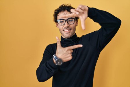 Téléchargez les photos : Homme hispanique debout sur fond jaune souriant faisant cadre avec les mains et les doigts avec le visage heureux. concept de créativité et photographie. - en image libre de droit
