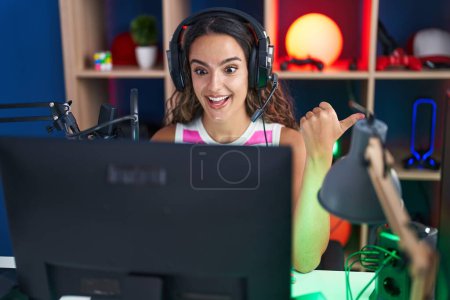 Téléchargez les photos : Jeune femme hispanique jouant à des jeux vidéo pointant le pouce vers le côté souriant heureux avec la bouche ouverte - en image libre de droit
