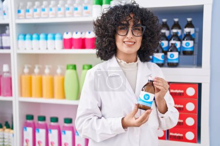 Foto de Joven farmacéutica de Oriente Medio sonriendo confiada sosteniendo la botella de medicamento en la farmacia - Imagen libre de derechos