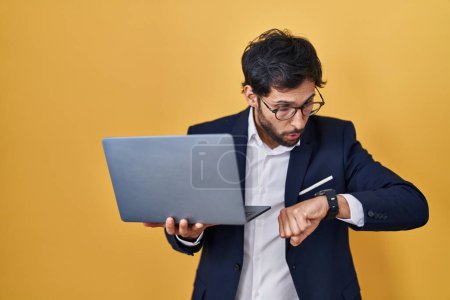 Téléchargez les photos : Bel homme latin travaillant à l'aide d'un ordinateur portable en regardant le temps de la montre inquiet, peur d'arriver en retard - en image libre de droit