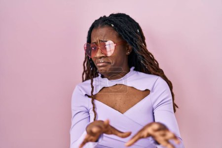 Téléchargez les photos : Femme africaine avec les cheveux tressés debout sur fond rose expression dégoûtée, mécontente et effrayée faisant visage dégoûtant parce que réaction d'aversion. les mains levées - en image libre de droit