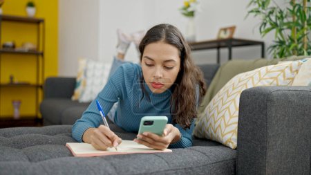 Photo for Young beautiful hispanic woman using smartphone writing on notebook lying on sofa at home - Royalty Free Image