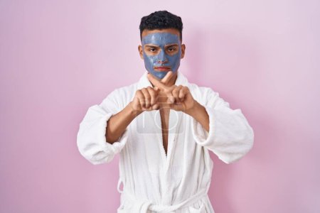 Foto de Joven hombre hispano usando mascarilla de belleza y expresión de rechazo de bata de baño cruzando dedos haciendo signo negativo - Imagen libre de derechos