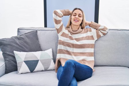 Foto de Young woman relaxed with hands on head sitting on sofa at home - Imagen libre de derechos