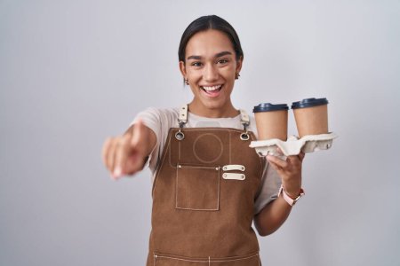 Foto de Mujer hispana joven vistiendo delantal de camarera profesional sosteniendo café señalándote a ti y a la cámara con los dedos, sonriendo positiva y alegre - Imagen libre de derechos