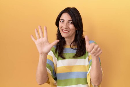 Foto de Mujer hispana de mediana edad de pie sobre fondo amarillo mostrando y señalando hacia arriba con los dedos número seis mientras sonríe confiado y feliz. - Imagen libre de derechos