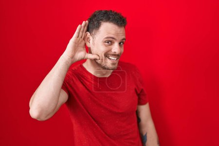 Photo for Young hispanic man standing over red background smiling with hand over ear listening an hearing to rumor or gossip. deafness concept. - Royalty Free Image
