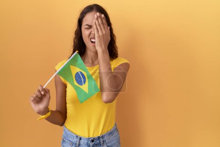 Foto de Mujer hispana joven sosteniendo bandera brasileña bostezando cansada cubriendo media cara, ojo y boca con la mano. cara duele en el dolor. - Imagen libre de derechos
