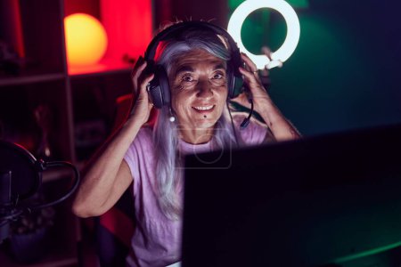 Foto de Streamer mujer de pelo gris de mediana edad sonriendo confiado sentado en la mesa en la sala de juegos - Imagen libre de derechos