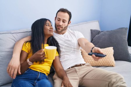Téléchargez les photos : Homme et femme interracial couple boire du café regarder la télévision à la maison - en image libre de droit