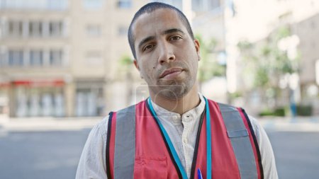 Téléchargez les photos : Jeune homme hispanique volontaire vêtu d'un gilet regardant sérieusement la rue - en image libre de droit