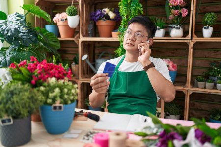 Photo for Young chinese man florist talking on smartphone holding credit card at flower shop - Royalty Free Image