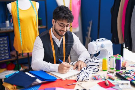 Téléchargez les photos : Young hispanic man tailor smiling confident drawing on notebook at sewing studio - en image libre de droit