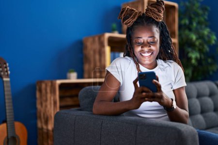 Foto de Mujer afroamericana usando teléfono inteligente sentado en el sofá en casa - Imagen libre de derechos
