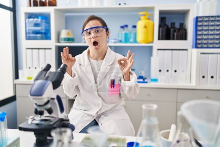 Photo for Hispanic girl with down syndrome working at scientist laboratory looking surprised and shocked doing ok approval symbol with fingers. crazy expression - Royalty Free Image