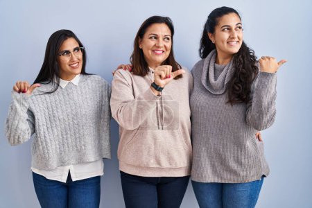 Photo for Mother and two daughters standing over blue background smiling with happy face looking and pointing to the side with thumb up. - Royalty Free Image