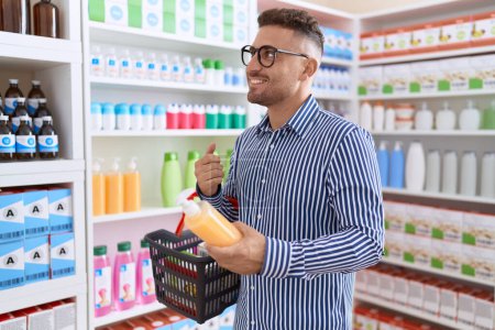 Téléchargez les photos : Jeune homme hispanique client souriant confiant tenant bouteille de shampooing et panier de marché à la pharmacie - en image libre de droit
