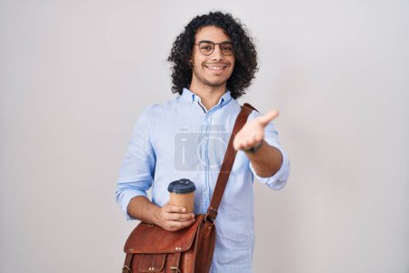 Foto de Hombre hispano con el pelo rizado bebiendo una taza de café para llevar sonriente amistoso ofreciendo apretón de manos como saludo y bienvenida. negocios exitosos. - Imagen libre de derechos