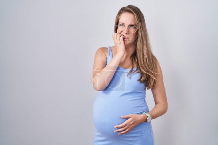 Téléchargez les photos : Jeune femme enceinte debout sur fond blanc regardant stressé et nerveux avec les mains sur les ongles mordants de la bouche. problème d'anxiété. - en image libre de droit