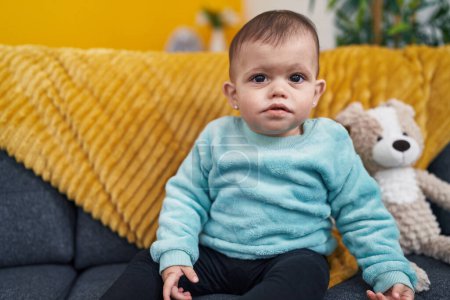 Téléchargez les photos : Adorable hispanic baby sitting on sofa with relaxed expression at home - en image libre de droit