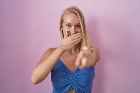 Photo for Young caucasian woman standing over pink background laughing at you, pointing finger to the camera with hand over mouth, shame expression - Royalty Free Image