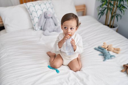 Photo for Adorable hispanic boy sitting on bed sucking bottle of water at bedroom - Royalty Free Image