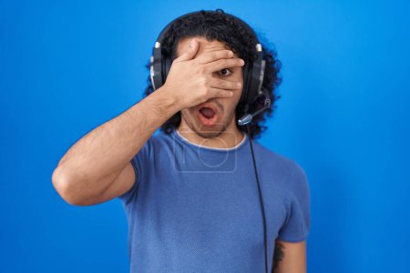 Photo for Hispanic man with curly hair listening to music using headphones peeking in shock covering face and eyes with hand, looking through fingers with embarrassed expression. - Royalty Free Image