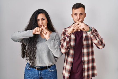 Foto de Joven pareja hispana de pie sobre fondo blanco expresión de rechazo cruzando dedos haciendo signo negativo - Imagen libre de derechos