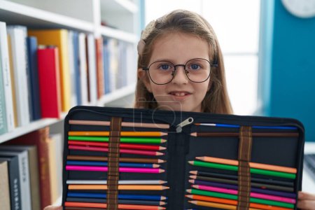 Téléchargez les photos : Adorable fille blonde étudiante souriante confiante tenant crayons de couleur cas à la salle de classe - en image libre de droit