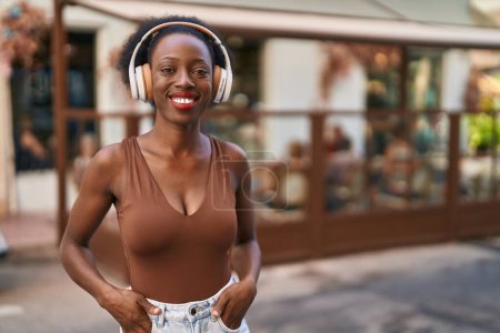 Téléchargez les photos : Femme africaine avec les cheveux bouclés en plein air à la ville portant des écouteurs collant la langue heureux avec une expression drôle. - en image libre de droit