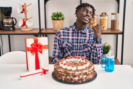 Téléchargez les photos : Homme africain avec dreadlocks célébrant anniversaire tenant grand gâteau au chocolat souriant avec visage heureux regardant et pointant vers le côté avec le pouce vers le haut. - en image libre de droit