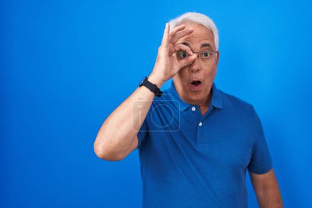 Téléchargez les photos : Homme du moyen âge avec les cheveux gris debout sur fond bleu faisant un geste correct choqué par le visage surpris, les yeux regardant à travers les doigts. expression incrédule. - en image libre de droit