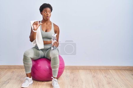 Foto de Mujer afroamericana con ropa deportiva sentada en una pelota de pilates sorprendida apuntando con el dedo de la mano hacia un lado, boca abierta expresión asombrada. - Imagen libre de derechos