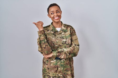 Téléchargez les photos : Belle femme afro-américaine portant l'uniforme de l'armée de camouflage souriant avec le visage heureux regardant et pointant vers le côté avec le pouce vers le haut. - en image libre de droit